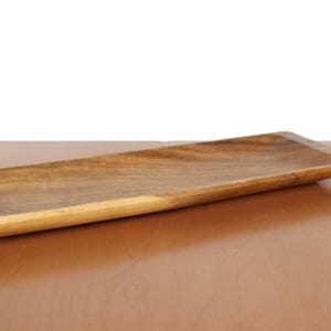 May include: A long, rectangular wooden serving tray with a natural wood finish. The tray is made of a single piece of wood and has a slightly curved shape.