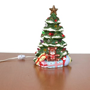 Könnte beinhalten: Ein Keramik-Weihnachtsbaum mit einem Teddybären und Geschenken an der Basis. Der Baum ist mit grünen, roten und weißen Ornamenten geschmückt und hat einen goldenen Stern oben. Der Baum ist mit weißen Lichtern beleuchtet und hat ein Kabel mit einem Schalter.