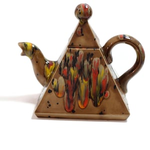 Puede incluir: Tetera de cerámica marrón con forma de pirámide y tapa a juego. La tetera tiene un asa y un pico, y está decorada con esmalte rojo, amarillo y naranja goteado. Una pieza única para servir té.