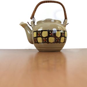 Pode incluir: Uma chaleira de cerâmica bege com uma alça tecida. A chaleira apresenta um padrão decorativo de quadrados amarelos com detalhes em vermelho e marrom. O bico e a tampa também são bege. A chaleira está sobre uma superfície marrom.
