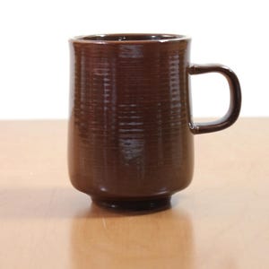 Könnte beinhalten: Eine braune Keramiktasse mit strukturierter Oberfläche und Henkel. Die Tasse hat eine zylindrische Form mit leicht ausgestelltem Boden. Die Tasse steht auf einer hellbraunen Oberfläche.