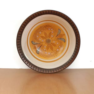 Puede incluir: Un cuenco redondo de cerámica con borde marrón e interior crema. El centro del cuenco presenta un diseño floral amarillo con detalles azules y grises. El cuenco está sobre una superficie marrón claro.