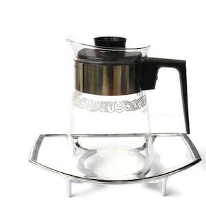 Puede incluir: Una cafetera de vidrio con una banda plateada y un asa negra se asienta sobre un soporte de metal plateado con tres patas. La cafetera tiene un diseño floral blanco.