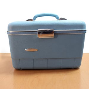 Könnte beinhalten: Ein vintage-blauer Kosmetikkoffer mit passendem Griff und silberfarbener Hardware. Der Koffer hat eine strukturierte Oberfläche, einen sicheren Verschluss und ein kleines ovales Emblem. Der Koffer steht auf einer Holzoberfläche.