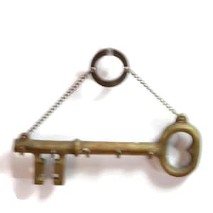 May include: A vintage-style key holder made of brass, featuring a large key design with multiple hooks for keys. The key is suspended by a chain from a circular ring. The key has a classic, antique aesthetic.