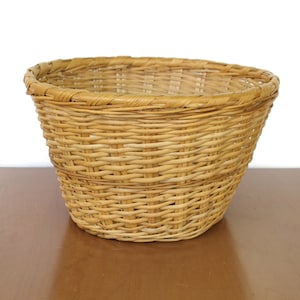May include: A woven wicker basket with a round, wide opening and a slightly tapered bottom. The basket is made of natural, light brown wicker.