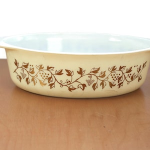 May include: Cream-coloured oval casserole dish with a gold grape and leaf design. The dish has a light blue interior and two handles. A vintage kitchenware item.