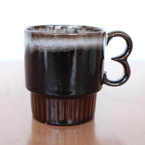 Könnte beinhalten: Eine braune und weiße Keramiktasse mit einem einzigartigen Doppelgriff. Die Tasse hat eine braune Glasur mit einem weißen Band um den oberen Rand.