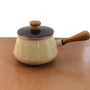 Puede incluir: Una cacerola beige vintage con tapa marrón oscuro y asa de madera. La olla tiene un patrón moteado y un borde plateado. El asa y el pomo de la tapa son de madera. Un artículo de cocina clásico.