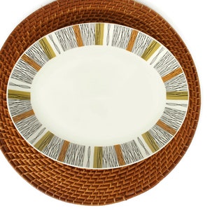 May include: A white oval serving platter with a brown, black, and gold geometric pattern around the rim. The platter is on a brown wicker charger.