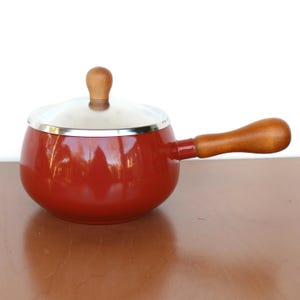 May include: Red enamel fondue pot with a wooden handle and a silver lid. The pot is sitting on a wooden surface.