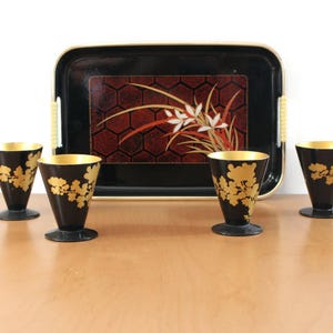 May include: A black lacquer serving tray with a red and gold floral design, accompanied by four black and gold sake cups. The cups have a gold interior and a floral pattern. The tray is approximately 14 inches long.