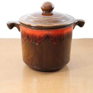 Puede incluir: Una cazuela de cerámica marrón con tapa. El plato tiene una base marrón oscuro con una banda roja vibrante cerca de la parte superior. Cuenta con dos asas laterales y un pomo en la tapa.