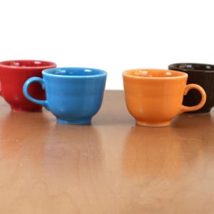 May include: Four colorful ceramic coffee cups in red, blue, orange, and brown are arranged on a light brown surface. Each cup has a rounded body and a curved handle. The cups are evenly spaced and set against a white background.