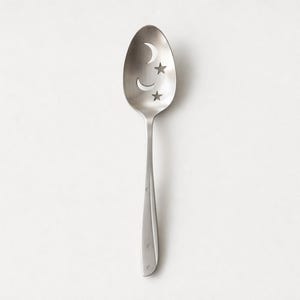 May include: A silver slotted spoon with a celestial design. The spoon's bowl features cut-out shapes of a crescent moon and two stars. The handle is long and slender, with a subtle design. The spoon is likely for kitchen use.