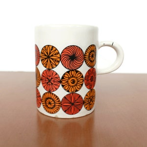 Puede incluir: Taza de cerámica blanca con un patrón repetitivo de diseños circulares en naranja y rojo. La taza tiene un asa curva. El diseño recuerda a los estilos retro o modernos de mediados de siglo. La taza está sobre un fondo neutro.