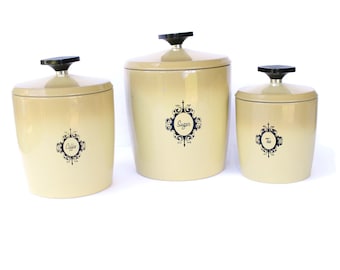 May include: Set of three vintage kitchen canisters in a pale yellow color with black accents. The canisters are labeled "Coffee", "Sugar", and "Tea" with a decorative design.