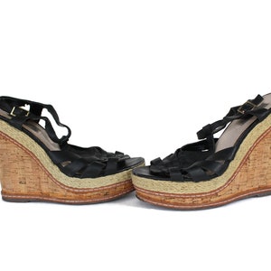 May include: A pair of black wedge sandals with a cork base and woven detailing. The sandals feature a strappy design with adjustable ankle straps and a platform sole. The wedge heel is approximately 4 inches high.