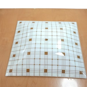 May include: Square, white and clear glass light fixture cover with a brown grid pattern and brown square accents. The cover has a single hole in the center and is slightly curved. The fixture is on a light brown surface.