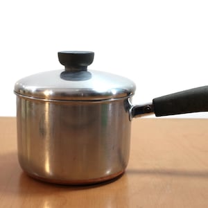 以下が含まれることがあります： 蓋と黒いハンドルが付いた、小さなステンレス製のソースパン。鍋底は銅製で、蓋には黒いノブが付いています。ハンドルには吊り下げ用のループが付いています。キッチン用品。