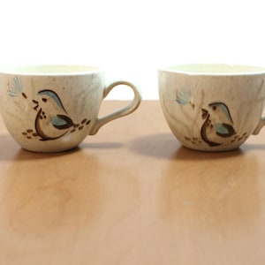 Könnte beinhalten: Zwei Keramik-Kaffeetassen mit Vogeldesign. Die Tassen sind beige mit einer braunen und blauen Vogelillustration. Jede Tasse hat einen gebogenen Henkel und eine weite Öffnung. Die Tassen stehen auf einer hellbraunen Oberfläche.