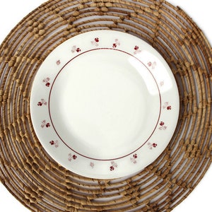 Könnte beinhalten: Ein weißer Keramikteller mit rotem und rosafarbenem Blumendesign auf einem geflochtenen braunen Untersetzer.