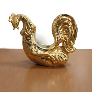 Könnte beinhalten: Eine goldfarbene Keramik-Hahn-Figur. Der dekorative Hahn hat einen detaillierten Schwanz und einen gebogenen Hals. Der Hahn ist ein Dekorationsartikel, wahrscheinlich für die Inneneinrichtung oder als Sammlerstück.