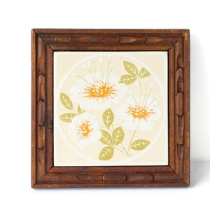 Può includere: Una piastrella di ceramica quadrata con un motivo floreale incorniciata da una cornice in legno marrone scuro. La piastrella presenta tre fiori bianchi con centri gialli e foglie verdi su uno sfondo crema.
