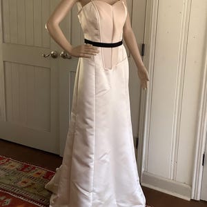 May include: A light peach-colored satin gown with a sweetheart neckline and spaghetti straps. The dress features a fitted bodice with a black belt and a flowing, floor-length skirt. The dress is displayed on a mannequin.
