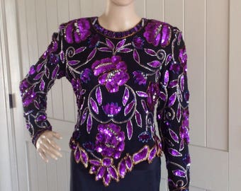 Könnte beinhalten: Ein langärmliges Paillettenoberteil mit einem floralen Design in Violett- und Goldtönen auf schwarzem Hintergrund.