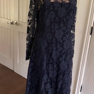 Op de afbeelding: Een zwarte kanten jurk met lange mouwen en een bloemenpatroon. De jurk heeft een transparante overlay en een gegolfde kanten zoom.