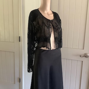 以下が含まれることがあります： 黒色の長袖ボレロジャケット。繊細なビーズとレースの装飾が施されています。黒のロングスカートと組み合わせています。マネキンに着せられており、デザインとフィット感を表現しています。