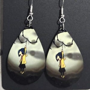 Peut inclure: Boucles d'oreilles en forme de goutte d'eau avec une illustration de dessin anim&eacute; d'une fille en manteau jaune suspendue &agrave; une branche sur fond de ciel nuageux. Les boucles d'oreilles ont des crochets d'oreille argent&eacute;s.