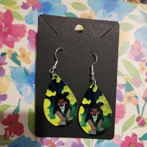 May include: Teardrop-shaped dangle earrings featuring a graphic illustration of a dark-maned lion with green eyes against a dark background with yellow smoke accents. The earrings are attached to a black rectangular display card with a top hole.
