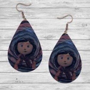 Peut inclure: Paire de boucles d'oreilles en forme de goutte d'eau avec une image de dessin anim&eacute; d'une fille aux cheveux noirs et aux yeux bleus. Les boucles d'oreilles ont un fond tourbillonnant bleu, rose et violet. Les crochets d'oreilles sont dor&eacute;s.