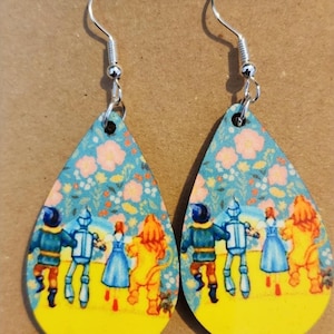 Puede incluir: Pendientes en forma de lágrima con ganchos plateados. Cada pendiente presenta una escena de El Mago de Oz, con Dorothy, el Hombre de Hojalata, el Espantapájaros y el León en un camino de baldosas amarillas, sobre un fondo floral.