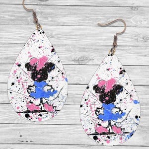 Op de afbeelding: Druppelvormige oorbellen met een witte achtergrond, bespat met roze, blauwe en zwarte verf. Elke oorbel heeft een cartoon-personageontwerp in zwart, roze en blauw, met een bronzen haak.