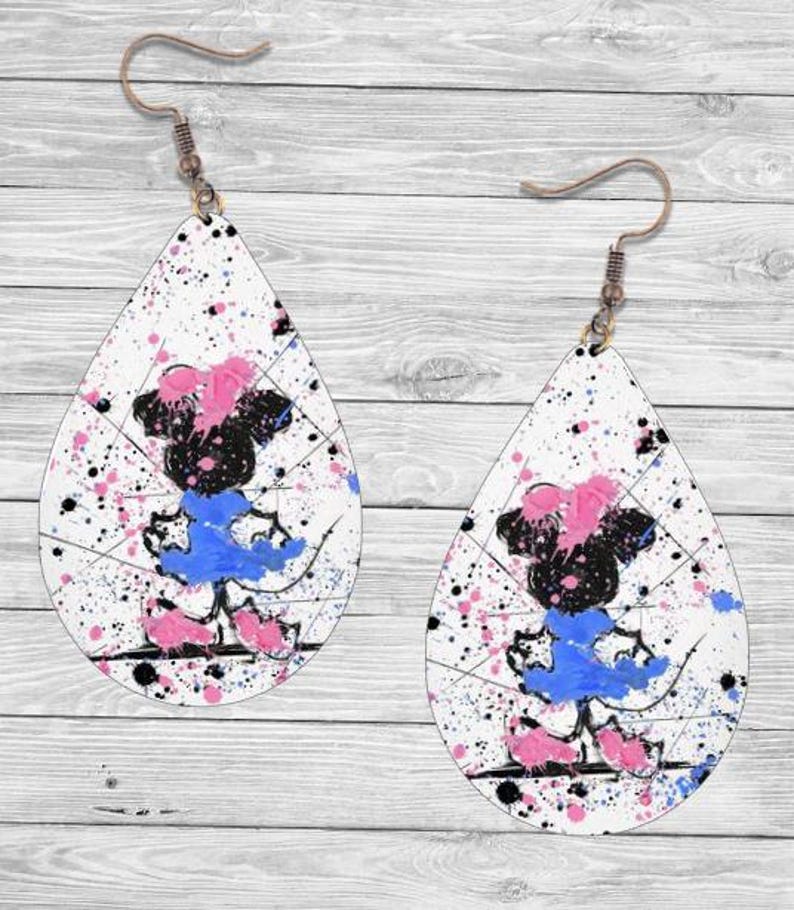Op de afbeelding: Druppelvormige oorbellen met een witte achtergrond, bespat met roze, blauwe en zwarte verf. Elke oorbel heeft een cartoon-personageontwerp in zwart, roze en blauw, met een bronzen haak.