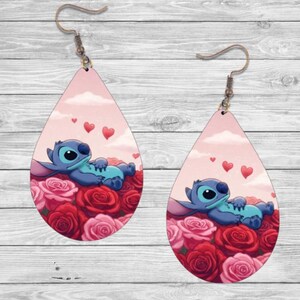 May include: A pair of teardrop-shaped earrings with a pink background and a blue cartoon character lying on a bed of red and pink roses. The character has a blue body, white eyes, and a pink nose. There are three small pink hearts floating above the character.