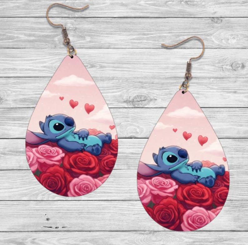May include: A pair of teardrop-shaped earrings with a pink background and a blue cartoon character lying on a bed of red and pink roses. The character has a blue body, white eyes, and a pink nose. There are three small pink hearts floating above the character.