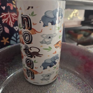 K&ouml;nnte beinhalten: Wei&szlig;er Reisebecher mit schwarzem Deckel, mit dem Namen "Nolan" in einer Schriftart im Dschungel-Stil. Der Becher ist mit Cartoon-Elefanten, Koalas, Tigern und Affen verziert. Das Design umfasst gr&uuml;ne Bl&auml;tter und orangefarbene Akzente.