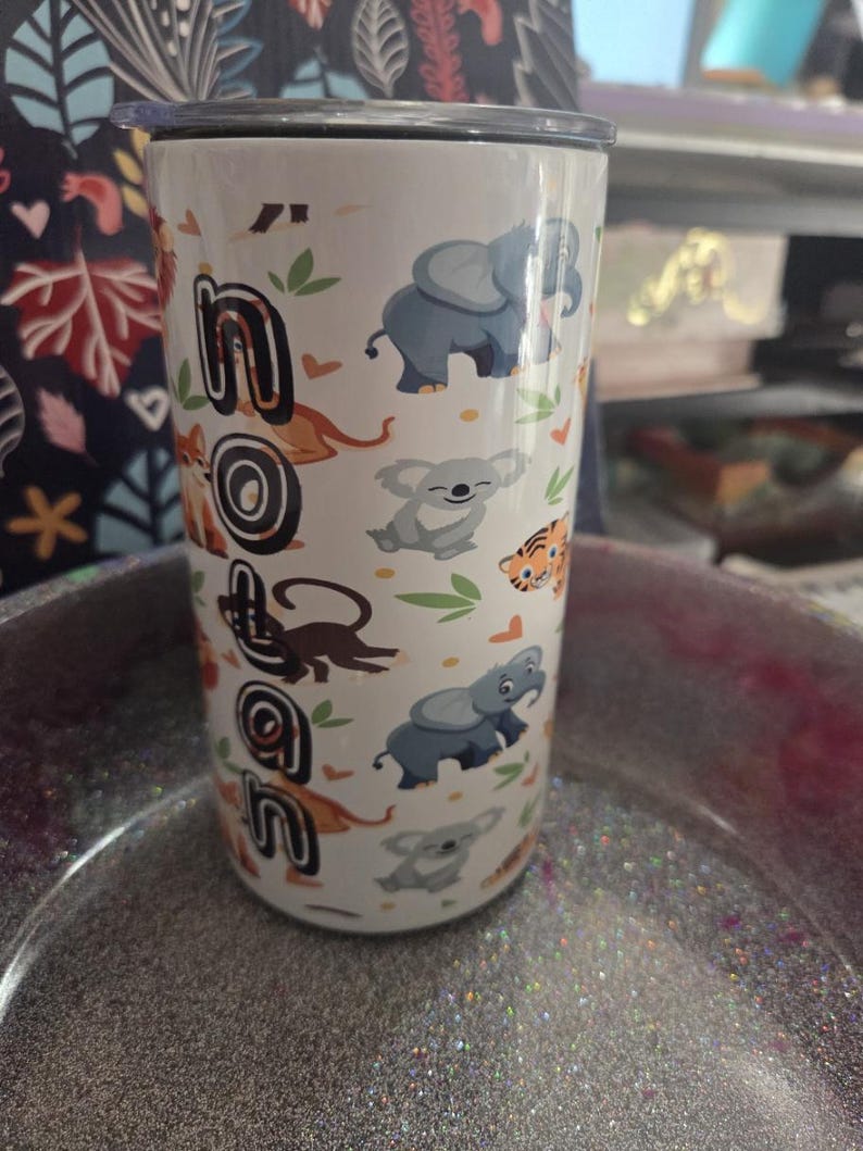 K&ouml;nnte beinhalten: Wei&szlig;er Reisebecher mit schwarzem Deckel, mit dem Namen "Nolan" in einer Schriftart im Dschungel-Stil. Der Becher ist mit Cartoon-Elefanten, Koalas, Tigern und Affen verziert. Das Design umfasst gr&uuml;ne Bl&auml;tter und orangefarbene Akzente.