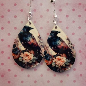 K&ouml;nnte beinhalten: Ohrringe in Tropfenform mit einem Rabenmuster. Der Rabe ist in Schwarz, Blau und Lila dargestellt, vor einem cremefarbenen Hintergrund. Das Blumenarrangement umfasst pfirsichfarbene, blaue und orangefarbene Rosen. Silberfarbene Haken.