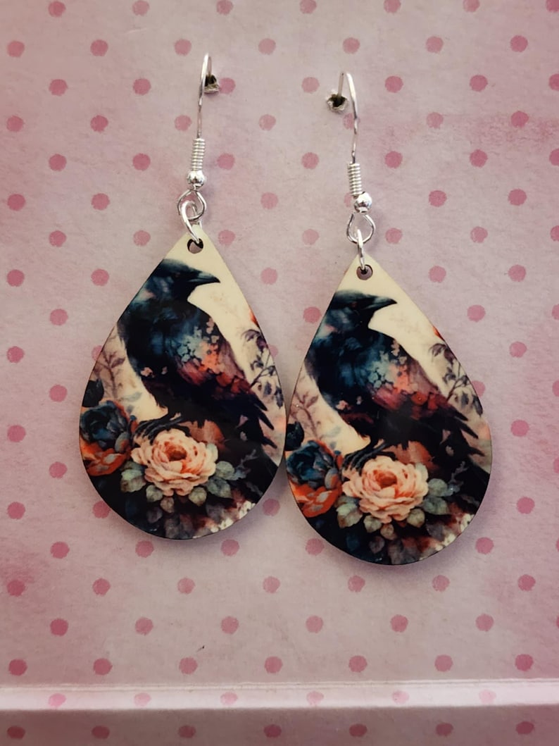 K&ouml;nnte beinhalten: Ohrringe in Tropfenform mit einem Rabenmuster. Der Rabe ist in Schwarz, Blau und Lila dargestellt, vor einem cremefarbenen Hintergrund. Das Blumenarrangement umfasst pfirsichfarbene, blaue und orangefarbene Rosen. Silberfarbene Haken.
