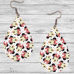 Op de afbeelding: Een paar druppelvormige oorbellen met een herhalend patroon van een stripfiguurgezicht met een rode strik op een witte achtergrond. De oorbellen hebben een bronzen haak.