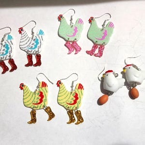 May include: A collection of whimsical chicken earrings. The earrings feature various chicken designs in white, green, and yellow, adorned with red combs and glittery boots. Some chickens have blue accents. The earrings are suspended from silver hooks.