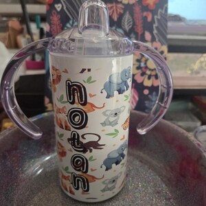 K&ouml;nnte beinhalten: Ein wei&szlig;er Trinkbecher mit transparentem Deckel und lila Griffen. Der Becher ist mit Zeichentricktieren und dem Namen "Nolan" in schwarzen Buchstaben verziert. Zu den Tieren geh&ouml;ren L&ouml;wen, Elefanten und Koalas.