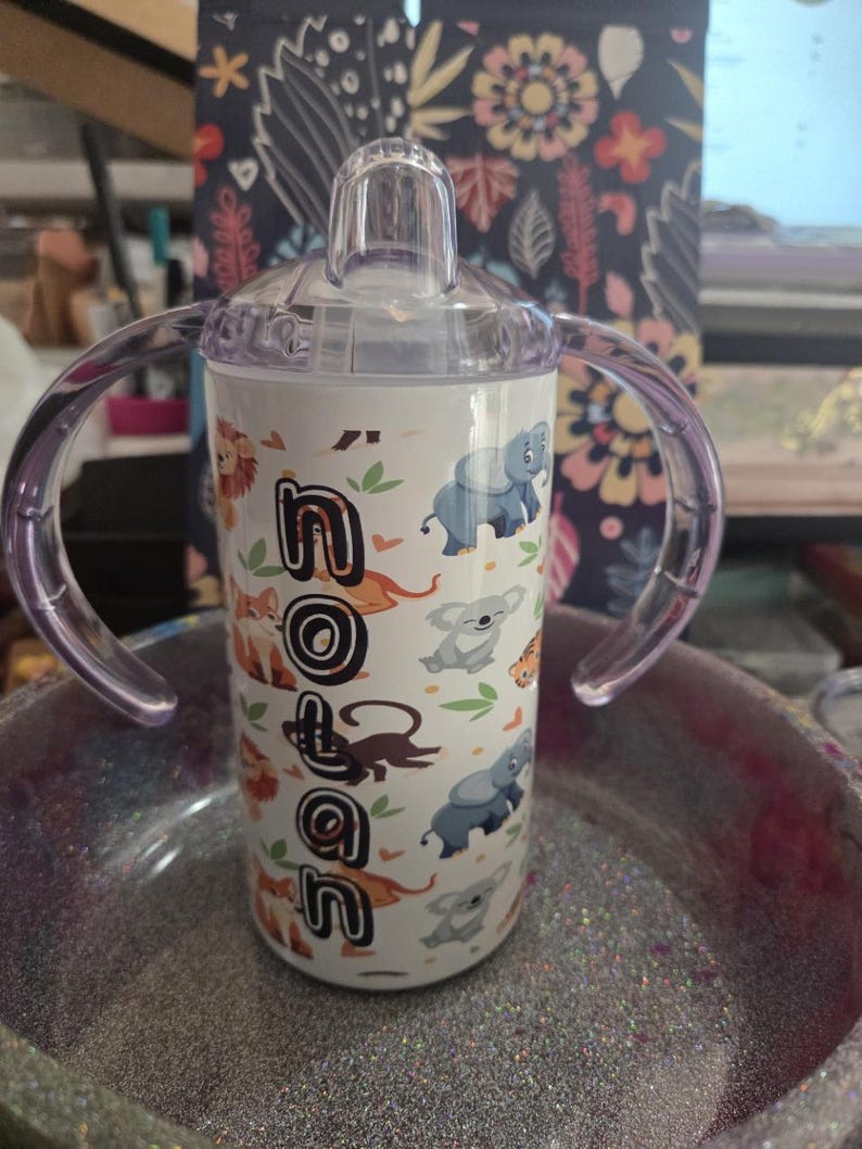 K&ouml;nnte beinhalten: Ein wei&szlig;er Trinkbecher mit transparentem Deckel und lila Griffen. Der Becher ist mit Zeichentricktieren und dem Namen "Nolan" in schwarzen Buchstaben verziert. Zu den Tieren geh&ouml;ren L&ouml;wen, Elefanten und Koalas.