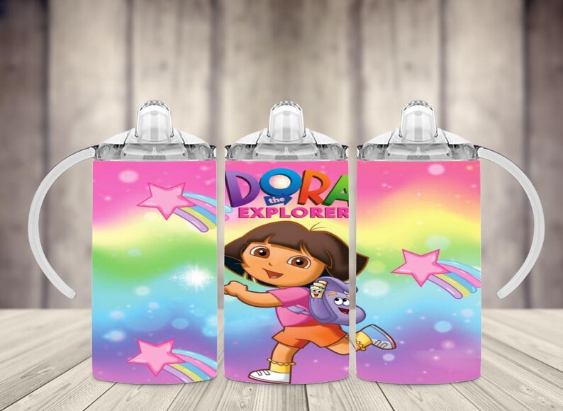 K&ouml;nnte beinhalten: Ein Trinkbecher f&uuml;r Kinder mit Dora der Entdeckerin. Der Becher hat einen rosa, blauen und gelben Hintergrund mit Sternen und Regenb&ouml;gen. Dora ist in der Mitte abgebildet, tr&auml;gt ein rosa Hemd und orangefarbene Shorts. Er hat einen wei&szlig;en Griff und Deckel.