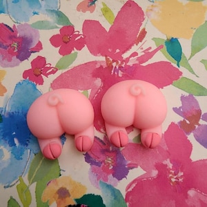 Peut inclure: Deux décorations roses en forme de derrière de cochon. Chacune a un corps rond, une queue en tire-bouchon et deux petites pattes. Elles sont posées sur un fond floral avec des fleurs roses, violettes et bleues.